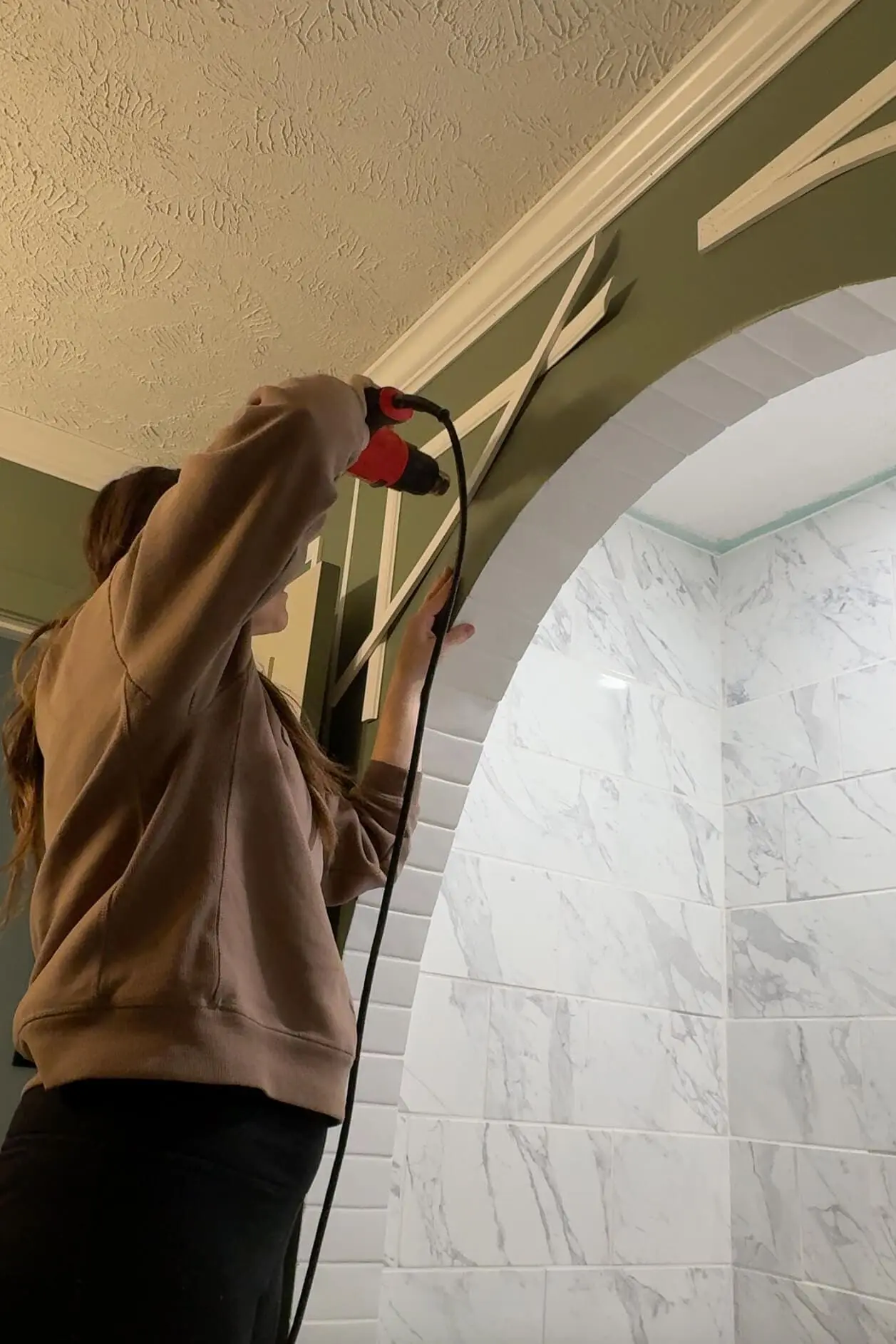Woman in a cozy brown sweatshirt using a heat gun to bend decorative molding around a green-painted archway in a DIY home improvement project. Bright bathroom tiles with a marble pattern peek through the arch for contrast.